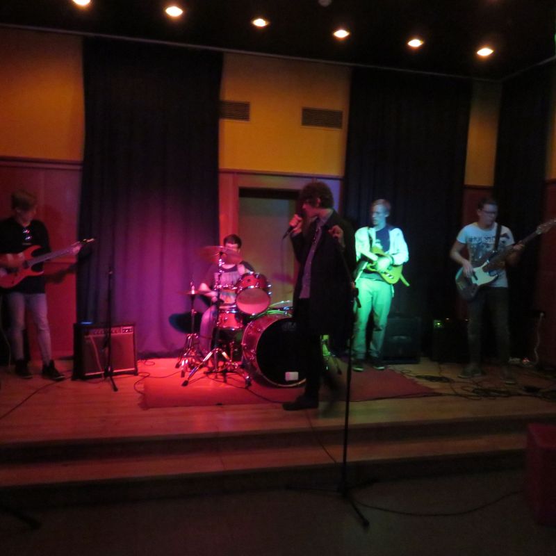 Auf einer Bühne stehen fünf Jugendliche und machen mit Schlagzeug, Gitarre, Bass und Gesang zusammen Musik.