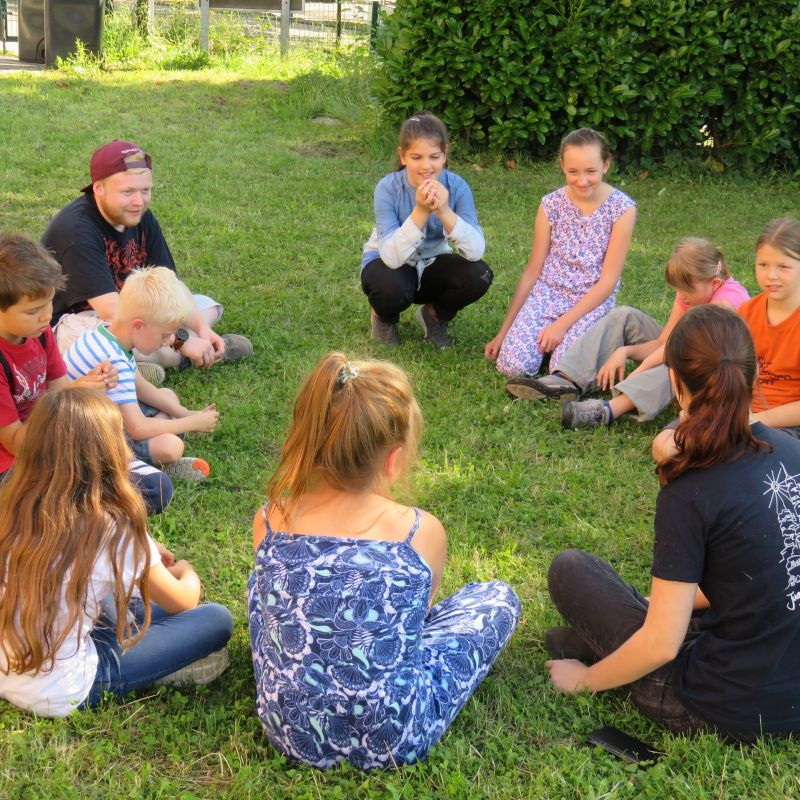 10 Kinder sitzen auf einer grünen wiese im Kreis und blicken sich gegenseitig an.