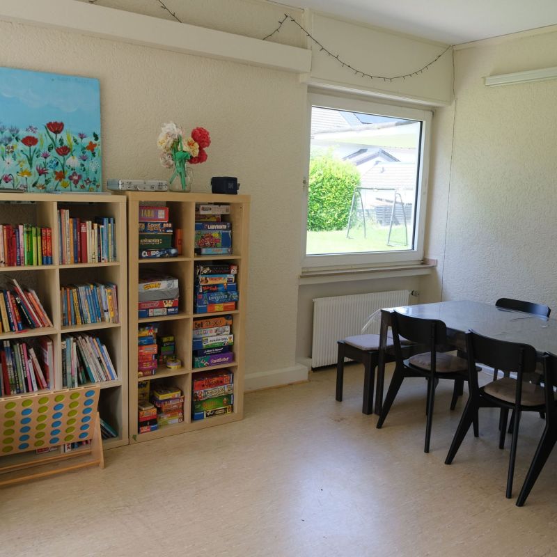 Offener Bereich mit Sitzgruppe und Tisch, einem Bücher- und Spieleregal.