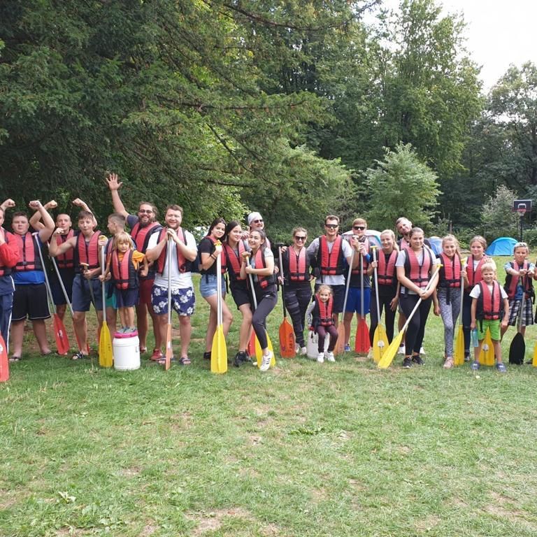 Jugendgruppe steht auf dem Rasen mit Schwimmweste und Paddeln und macht und Gruppenfoto vor dem Kanufahren.