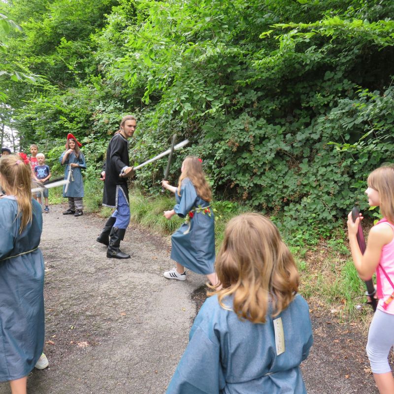 Kinder sind in der Natur auf Schnitzeljagd und treten spielerisch in Holzschwertkämpfen gegeneinander an.