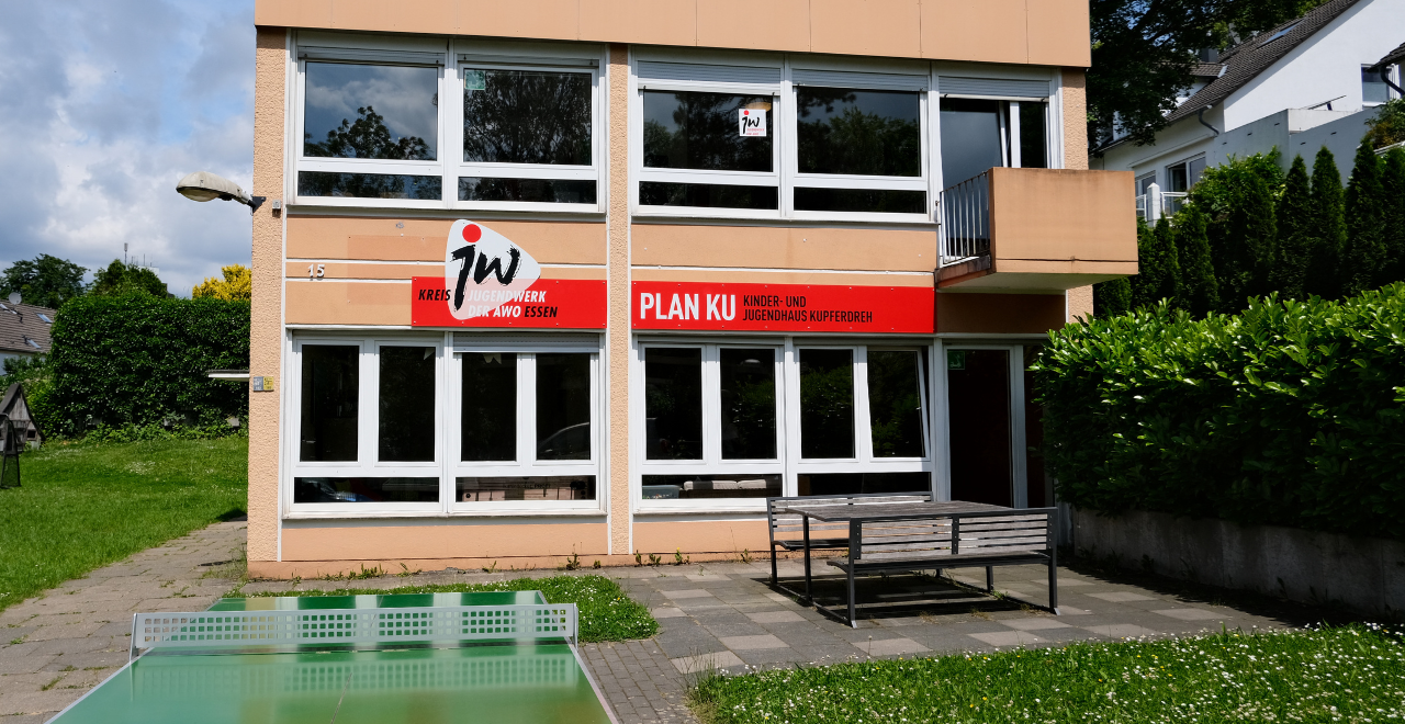 Foto vom Kinder- und Jugendhaus Plan KU. Es ist orange, hat zwei Etagen und davor ist eine Sitzgruppe, Rasen und eine Tischtennisplatte.