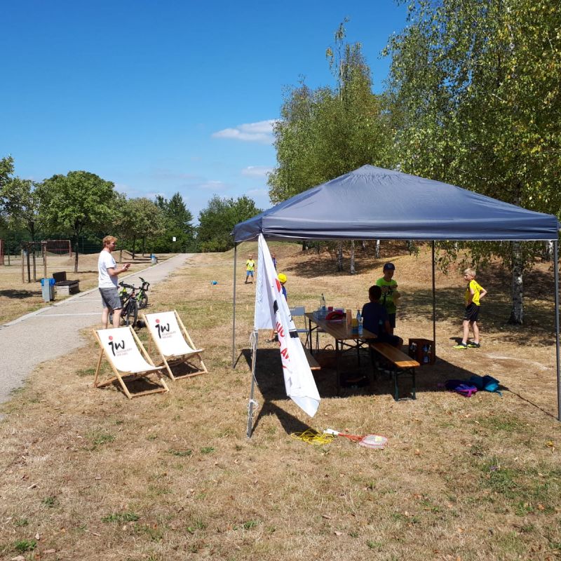 Auf einer Wiese steht ein Pavillon mit einer Jugendwerksfahne davor und zwei Liegestühlen. Auf der Wiese stehen Bäume und es laufen Kinder umher und spielen.