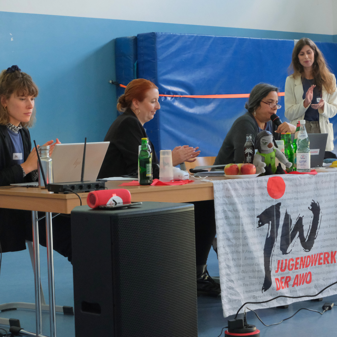 Die Konferenzleitung und eine vortragende Person sitzen vorne am Pult und halten eine Rede.