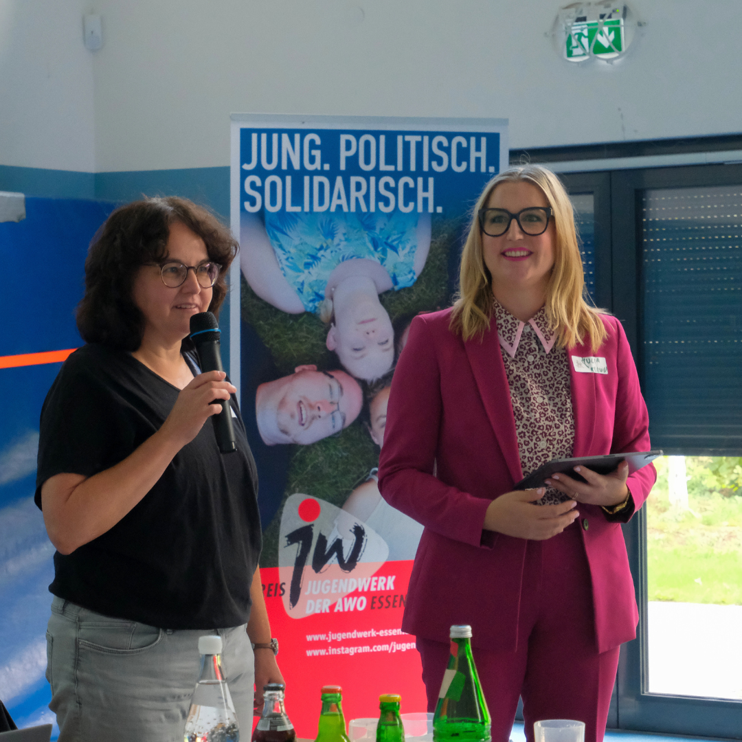 Die Moderation der Konferenz und eine Gastrednerin stehen vorne am Pult und sprechen zum Publikum.